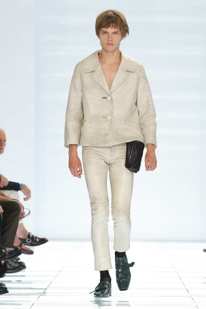A Prada model walks the spring 2027 runway in a fitted off-white leather peplum jacket with wide notch lapels over slim off-white leather trousers, holding a small black leather clutch and wearing dark green strap shoes