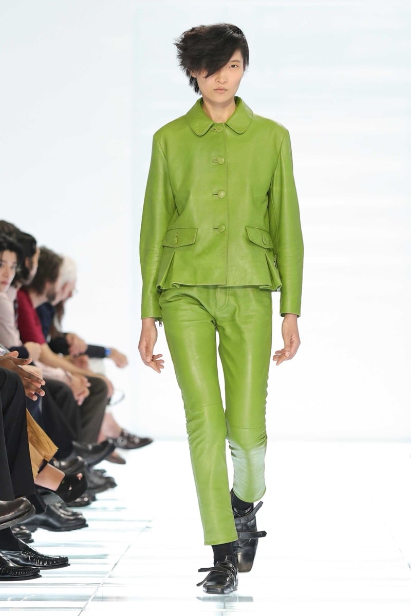 A Prada model walks the spring 2027 runway in a bright lime green leather peter pan collar jacket with covered buttons and matching lime green leather trousers, paired with black multi-strap leather shoes
