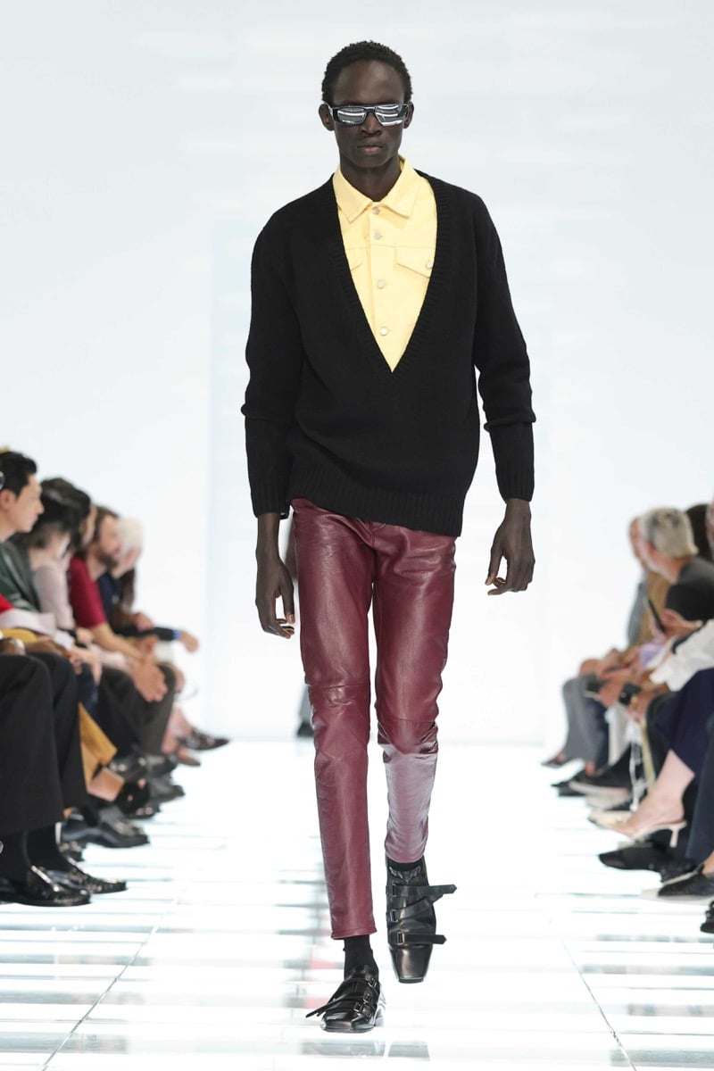 A Prada model walks the spring 2027 runway in narrow sunglasses, a black wool V-neck sweater over a butter yellow denim shirt, paired with slim burgundy leather trousers and black strap shoes