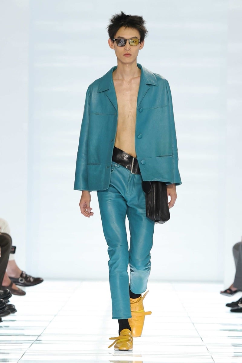A Prada model walks the spring 2027 runway in tinted sport sunglasses and an unbuttoned teal leather blazer over bare chest, paired with matching teal leather trousers, a black leather belt and pouch, and yellow strap shoes