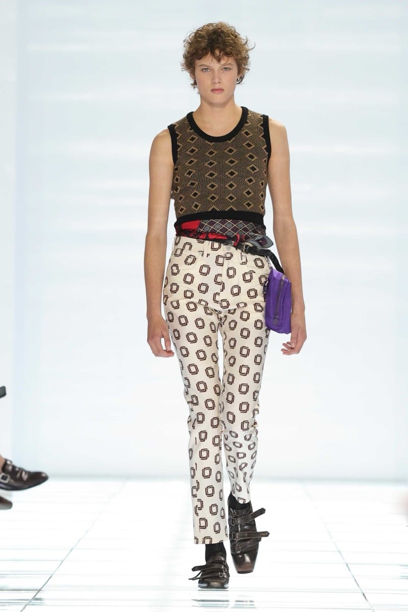 A Prada model walks the spring 2027 runway in a black and tan diamond-knit tank top, paired with cream geometric-print trousers, a red foulard tied at the waist, and a purple Prada nylon belt pouch