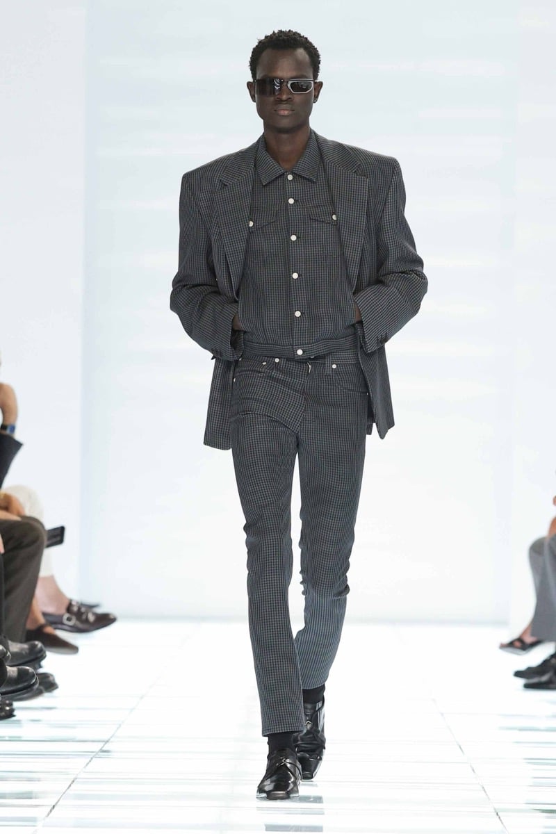 A Prada model walks the spring 2027 runway in narrow black sport sunglasses and a charcoal mini-gingham suit worn over a matching charcoal gingham shirt, finished with black leather loafers