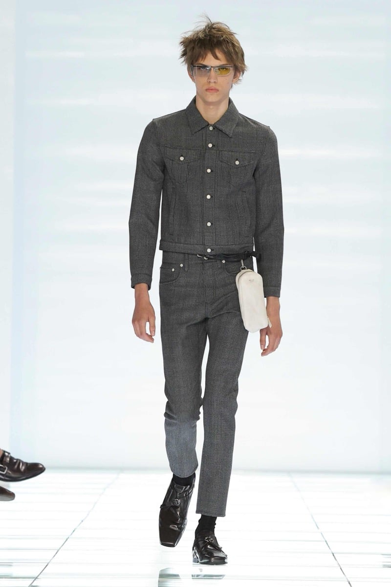A Prada model walks the spring 2027 runway in tinted yellow sunglasses and a cropped gray Prince of Wales check denim-style jacket with matching slim check trousers, carrying a small ivory Prada leather pouch