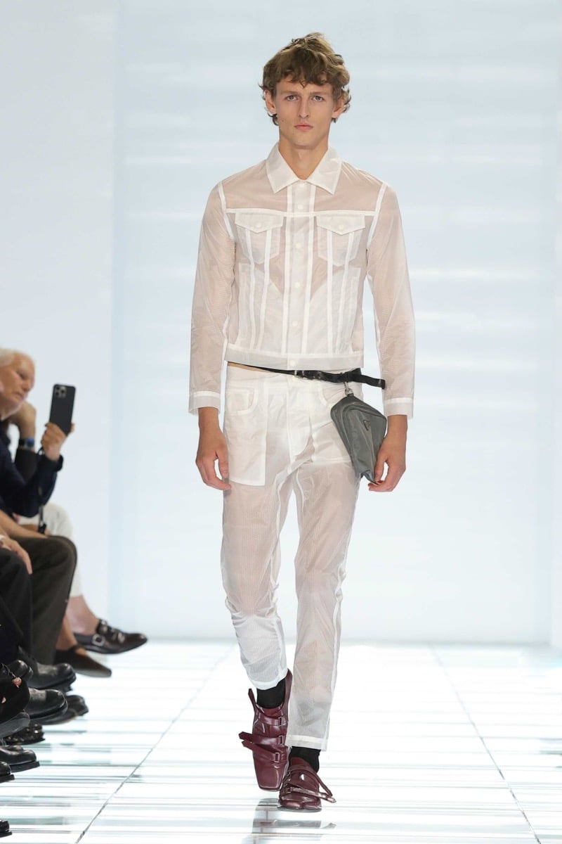 A Prada model walks the spring 2027 runway in a sheer white mesh trucker jacket and matching sheer mesh trousers worn over briefs, with a black belt holding a gray Prada nylon pouch and burgundy strap shoes