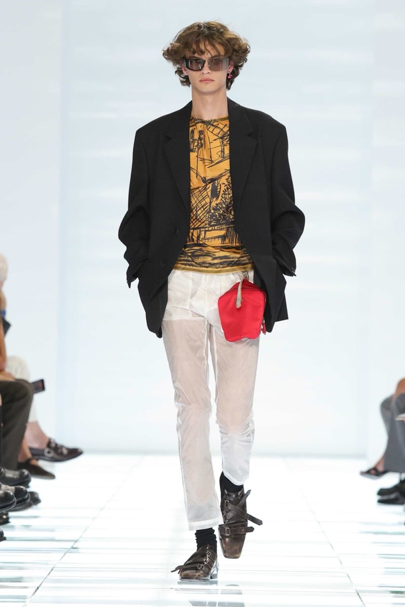A Prada model walks the spring 2027 runway in narrow rectangular sunglasses and an oversized black blazer over a mustard yellow printed T-shirt, paired with sheer white trousers, a red Prada nylon pouch, and brown multi-strap shoes