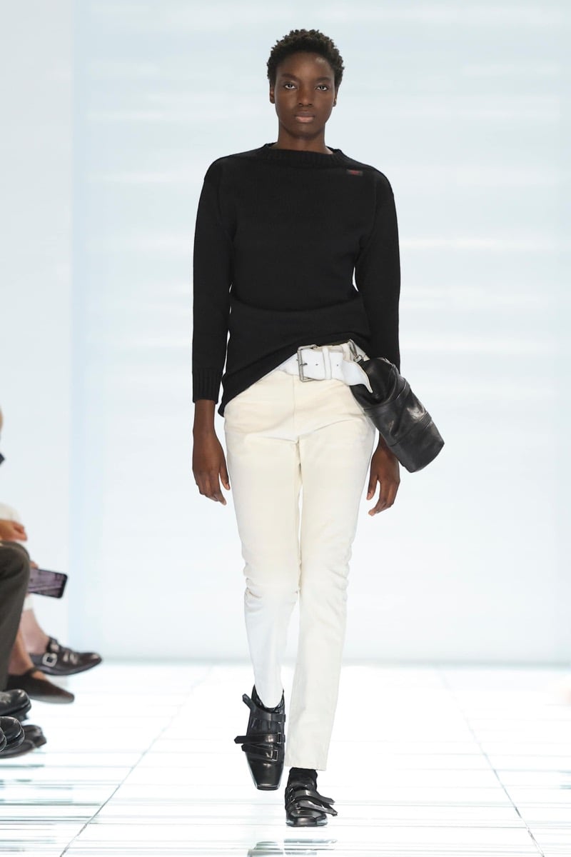 A Prada model walks the spring 2027 runway in a black fine-knit boatneck sweater layered over ivory leather trousers cinched with a wide white belt, holding a black leather pouch and wearing black strap shoes