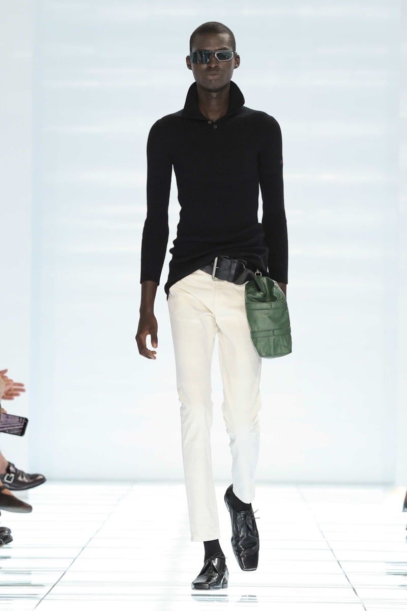 A Prada model walks the spring 2027 runway in narrow black sport sunglasses, a slim black ribbed turtleneck sweater, and ivory leather trousers with a black belt holding a forest green leather pouch, finished with black leather shoes