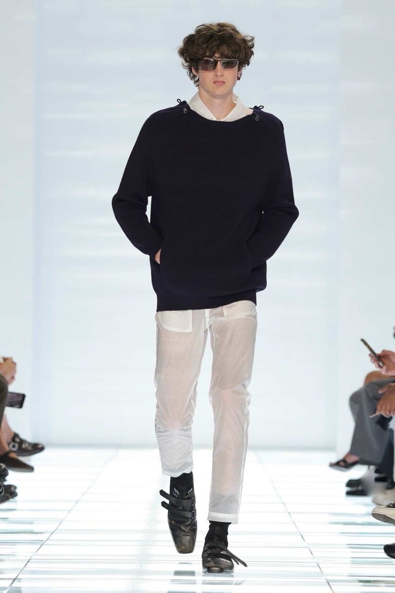 A Prada model walks the spring 2027 runway in shield sunglasses and a navy ribbed crewneck sweater with shoulder buttons layered over a white shirt, paired with sheer white mesh trousers and black strap shoes
