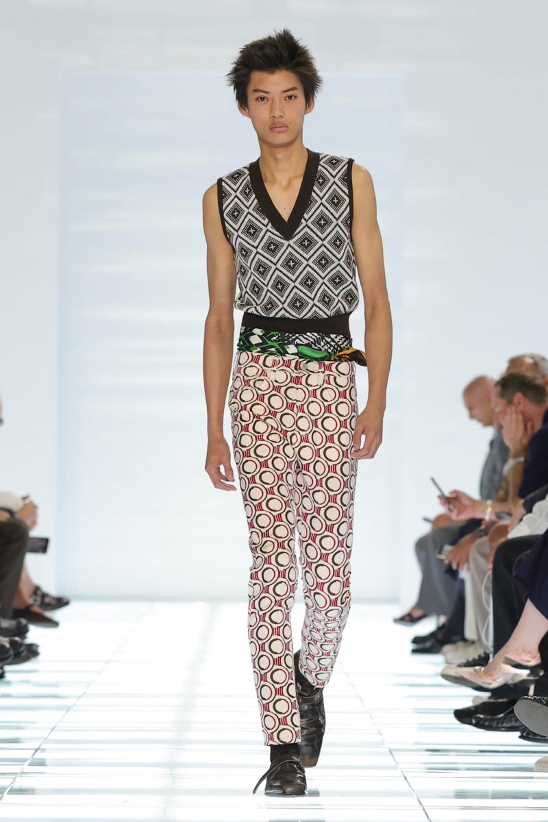 A Prada model walks the spring 2027 runway in a black and ivory diamond-knit V-neck sleeveless vest, paired with cream and burgundy circle-print trousers, a printed silk scarf knotted at the waist, and black strap shoes