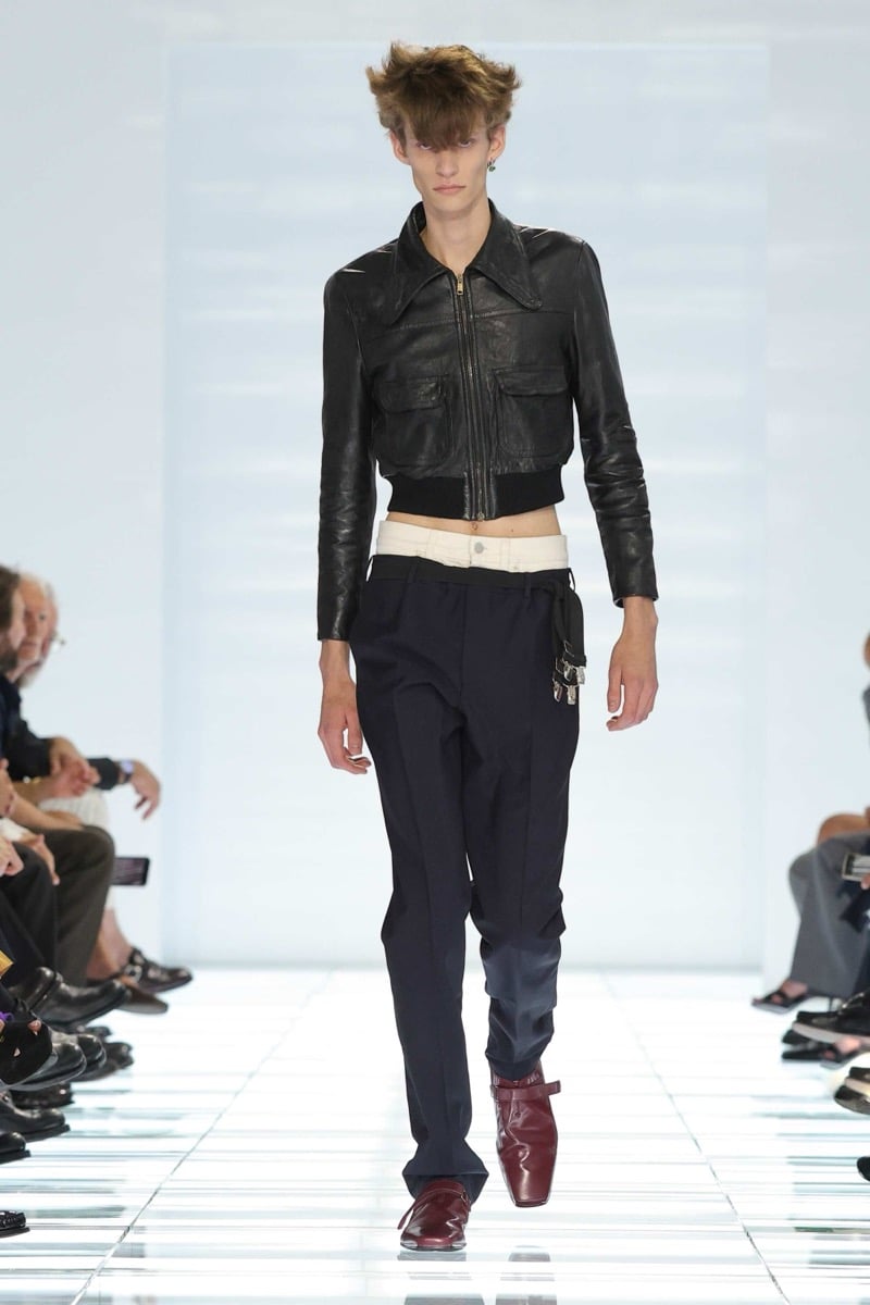A Prada model walks the spring 2027 runway in a cropped black leather bomber jacket with twin flap pockets over navy pleated trousers layered above an ivory denim waistband, finished with burgundy strap shoes