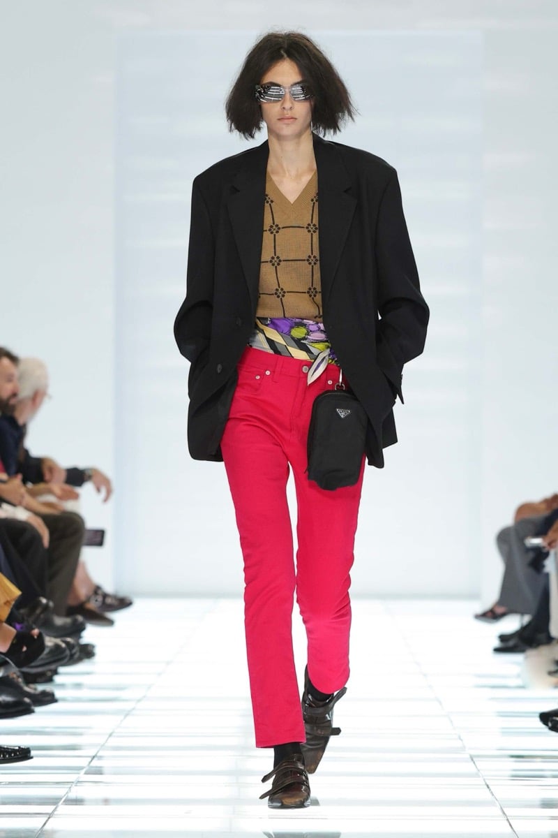 A Prada model walks the spring 2027 runway in narrow rectangular sunglasses and an oversized black blazer over a tan grid-knit vest, paired with bright fuchsia slim jeans, a striped silk scarf at the waist, and a black Prada triangle-logo pouch