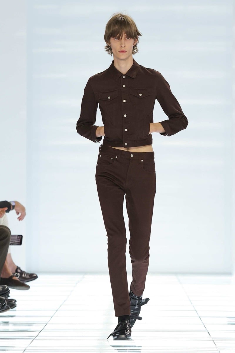 A Prada model walks the spring 2027 runway in a cropped dark brown denim jacket and matching slim dark brown jeans, finished with black multi-strap leather shoes