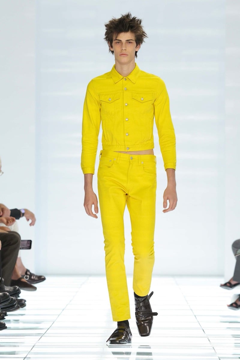 A Prada model walks the spring 2027 runway in a cropped bright yellow denim jacket and matching slim yellow jeans, finished with black multi-strap leather shoes