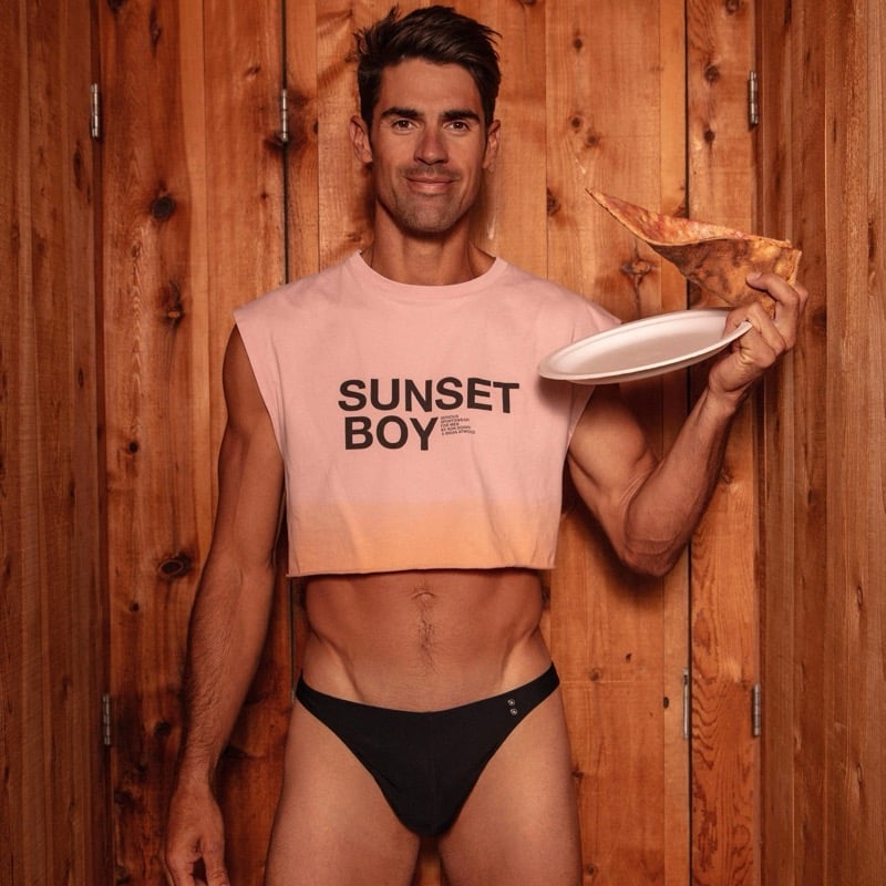 Chad White wears the Sunset Boy cropped tank in pink and holds a slice of pizza on a paper plate against a wood-paneled wall