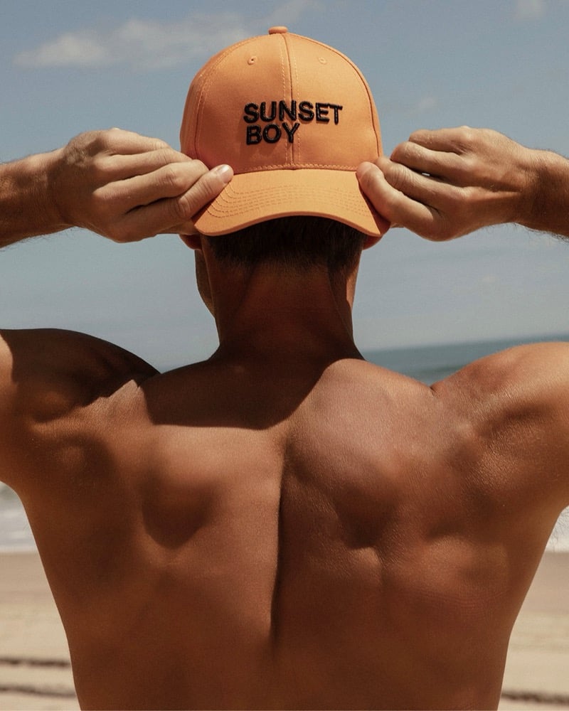 Back view of Chad White adjusting an orange Sunset Boy embroidered cap on the beach