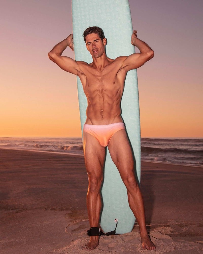 Chad White poses on the beach at sunset holding a surfboard upright behind him, wearing a sunset-gradient swim brief