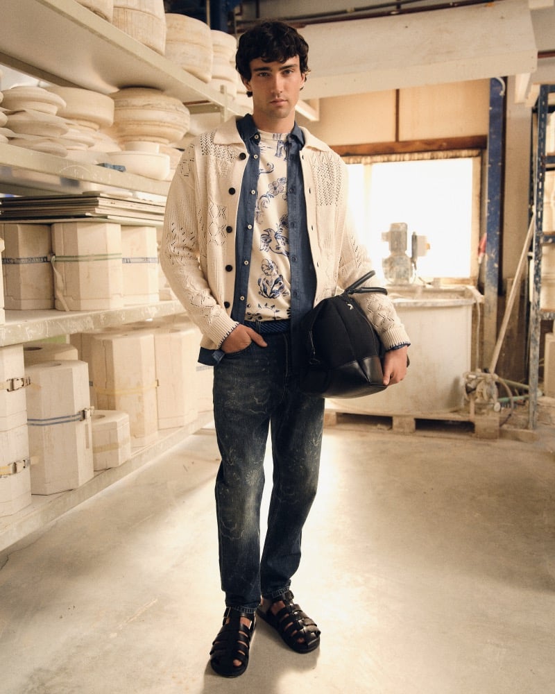 Joe Barnes standing in a Royal Delft pottery workshop lined with porcelain molds, wearing a cream openwork knit cardigan layered over a navy denim shirt and a Delft Blue floral printed tee, dark indigo jeans, and brown leather fisherman sandals
