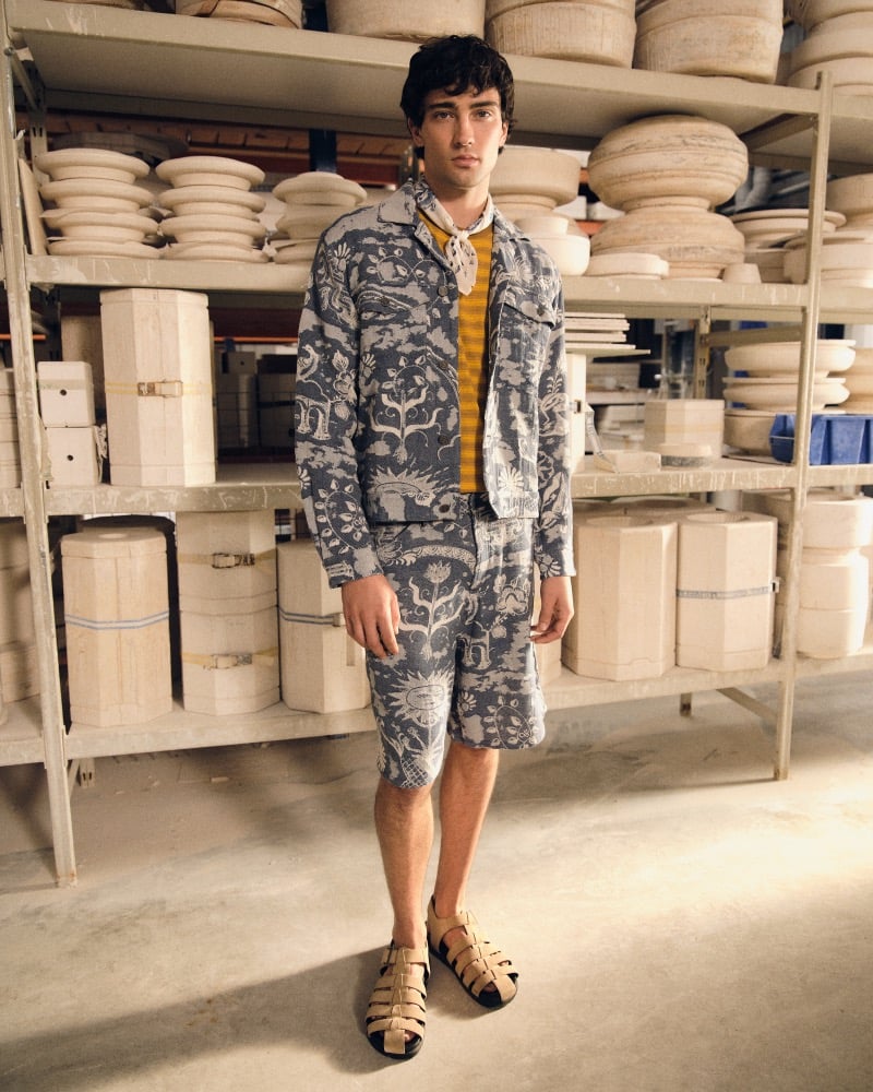 Joe Barnes inside the Royal Delft pottery studio in a matching Delft Blue jacquard chore jacket and walking shorts set worn over a yellow striped tee, with woven tan and black fisherman sandals