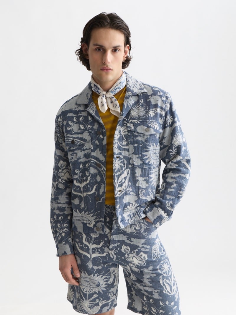 Scotch & Soda x Royal Delft jacquard chore jacket and matching shorts in indigo and ivory Delft Blue motifs, layered over a yellow striped tee and a Delft Blue printed bandana tied at the neck