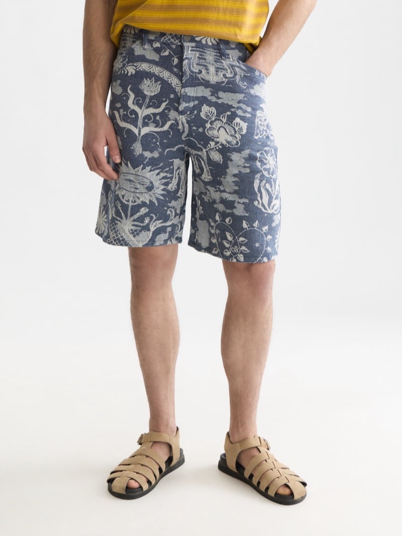 Scotch & Soda x Royal Delft jacquard shorts with sun, floral, and pastoral motifs in ivory on indigo, worn with a yellow striped tee and tan suede fisherman sandals