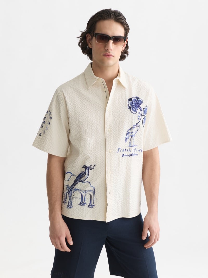 seersucker short-sleeve camp shirt printed with Delft Blue motifs of a giraffe under an archway, a peacock, and a flower, paired with navy trousers and gradient brown rectangular sunglasses