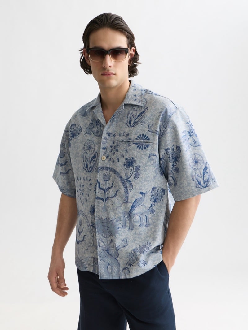 boxy short-sleeve camp shirt printed all over with pale blue Delft Blue ornament featuring birds, flowers, and scrollwork, worn open at the collar with navy trousers and rectangular sunglasses