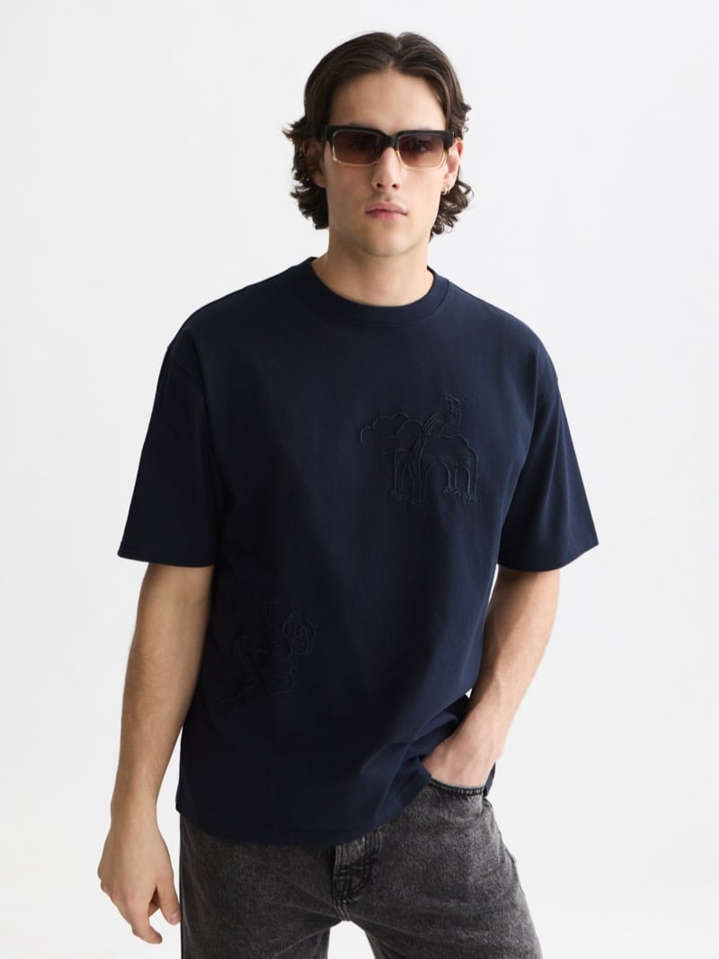 oversized navy cotton t-shirt with tonal embroidery of a giraffe under a Delft Blue archway and a small floral motif, worn with washed black denim and brown rectangular sunglasses