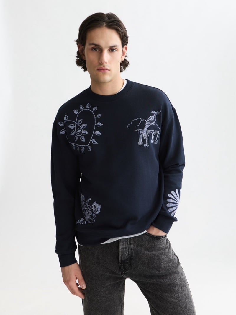 navy crewneck sweatshirt embroidered with pale blue Delft Blue motifs of a peacock under an archway, a sun-rayed heart, and floral scrollwork, layered over a white tee with charcoal denim
