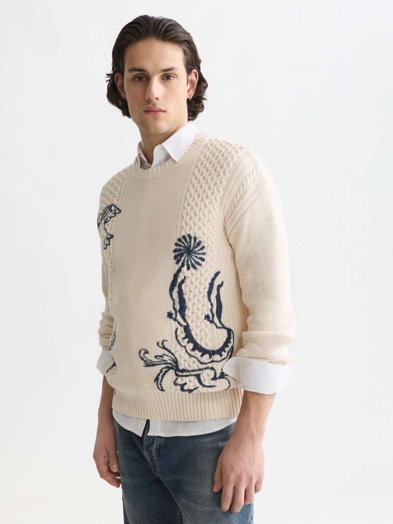 cream textured cable-knit crewneck sweater intarsia-knit with navy Delft Blue floral and scroll motifs, layered over a white oxford shirt with rolled cuffs and worn with mid-blue jeans