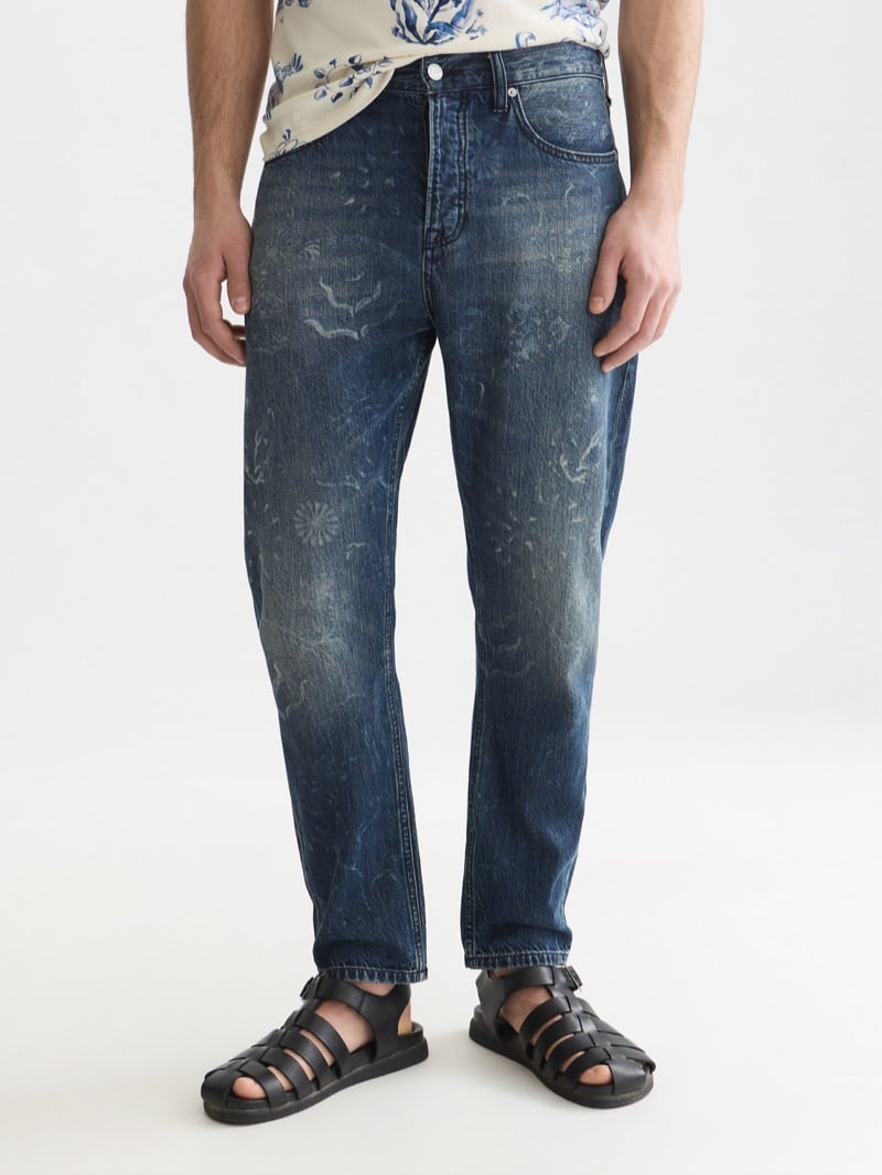 mid-wash blue tapered jeans printed with faded Delft Blue motifs, paired with black leather fisherman sandals and an untucked floral printed shirt