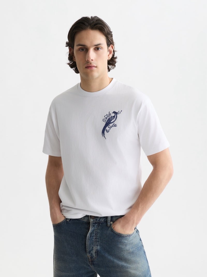 white cotton t-shirt embroidered with the Scotch & Soda peacock script logo in navy at the left chest, tucked into mid-wash blue jeans