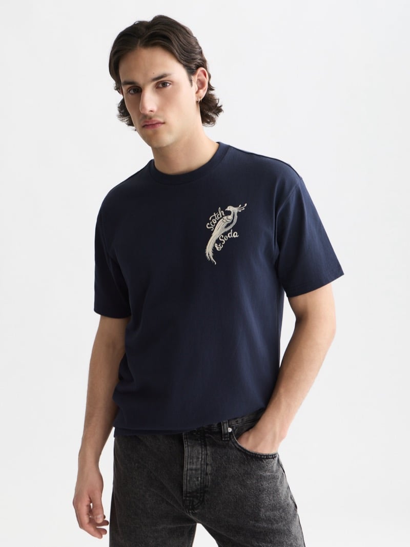 navy boxy-fit t-shirt embroidered with the ivory Scotch & Soda peacock script logo at the left chest, paired with washed black jeans