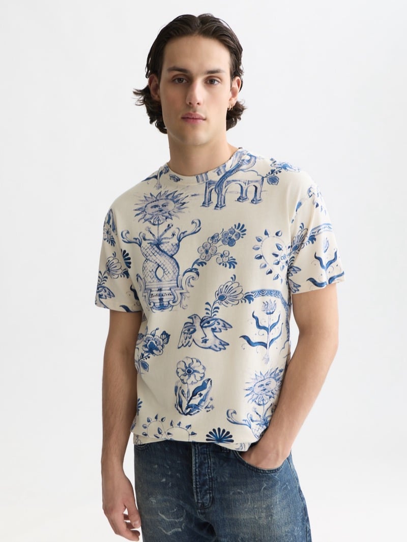 cream t-shirt printed all over with Delft Blue ornament including a sun, a giraffe under an archway, birds, and floral motifs, worn with mid-blue jeans flecked with the same pattern