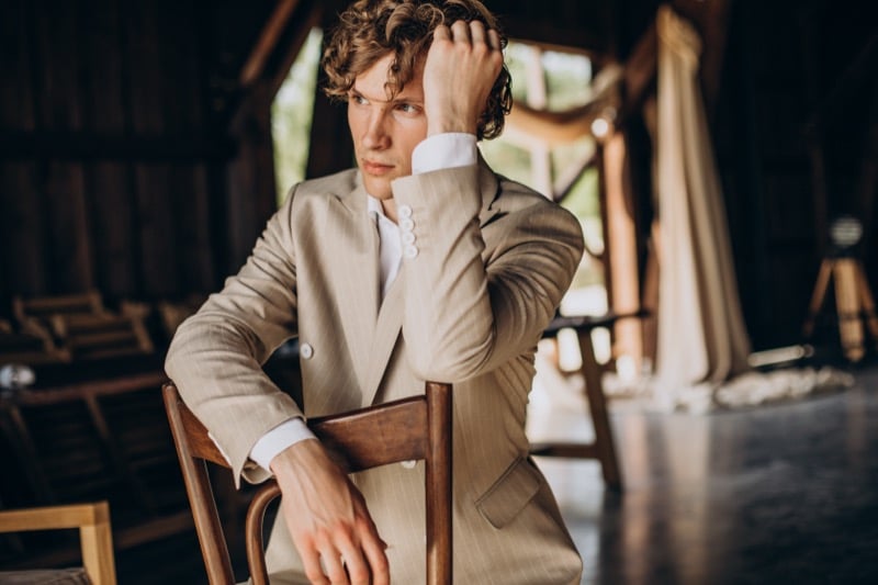 Curly-haired man in a beige bespoke summer suit and a buttoned dress shirt seated on a wooden chair inside a barn-style venue, the soft shoulder line and clean lapel of a lightweight summer cloth