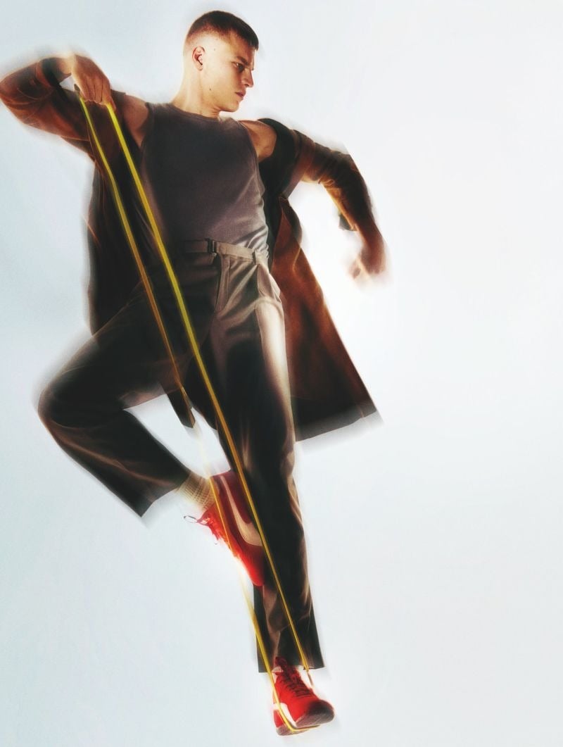 Timo Pan in a gray ribbed tank tucked into brown belted trousers and a brown coat slung over his shoulders, anchoring a yellow resistance band between his hands and a raised foot in a red running sneaker, blurred motion against a white backdrop
