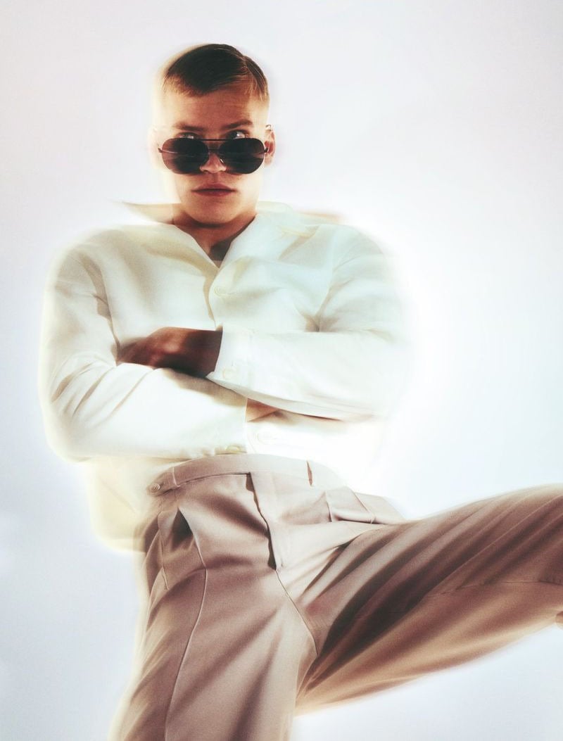 Timo Pan with arms crossed and oval rimless sunglasses in a linen shirt jacket and pale stone pleated wide-leg trousers, photographed from a low angle against a white backdrop with a slight motion blur