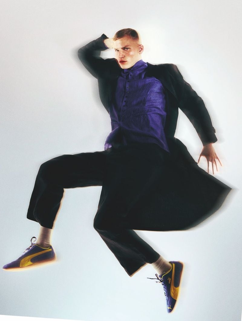 Timo Pan suspended mid-leap with one hand to his head, dressed in a dark coat over a purple linen long shirt and black trousers, white tube socks and navy Puma suede sneakers with yellow stripes