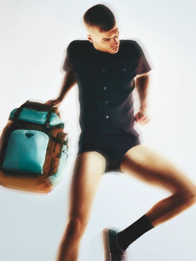 Timo Pan in a short-sleeve snap-front utility playsuit with built-in shorts, gripping the strap of a mint and olive Prada nylon backpack against a white floor, one bent leg raised into the frame