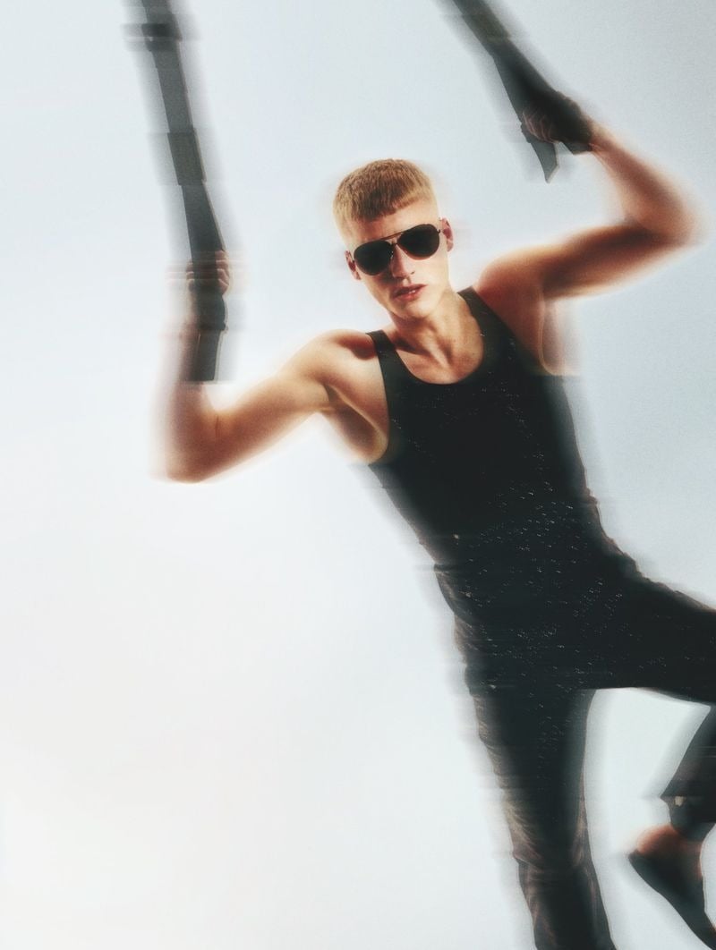 Timo Pan with a bleached crop and aviator sunglasses, dressed in a fitted black tank and dark trousers, grips two black suspension straps with motion-blur trails against a soft white backdrop.