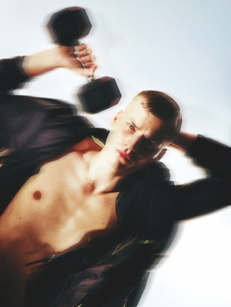 Overhead view of Timo Pan reclining bare-chested in an open dark zip jacket, slick-back blond hair against a white floor, lifting a black hex dumbbell above his head with one hand