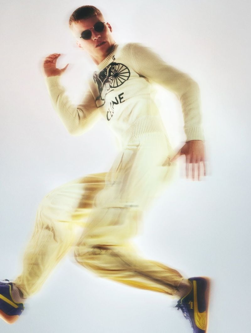 Timo Pan caught mid-stride in a Celine intarsia knit sweater printed with a vintage penny-farthing bicycle motif over cream drawstring trousers and navy Puma suede sneakers with yellow stripes, round sunglasses and a motion-blur trail across a white backdrop