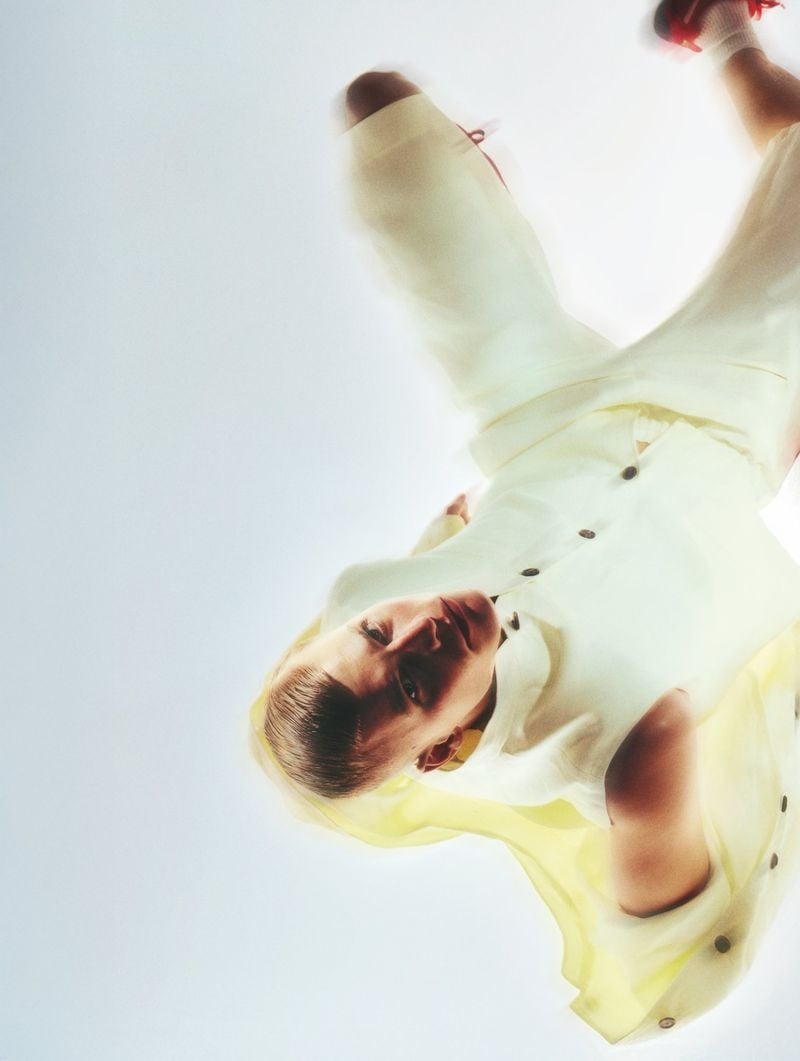 Inverted overhead shot of Timo Pan in a sheer pale yellow shirt with buttons and white tailored trousers, white tube socks and red running sneakers raised into the frame against a soft white backdrop