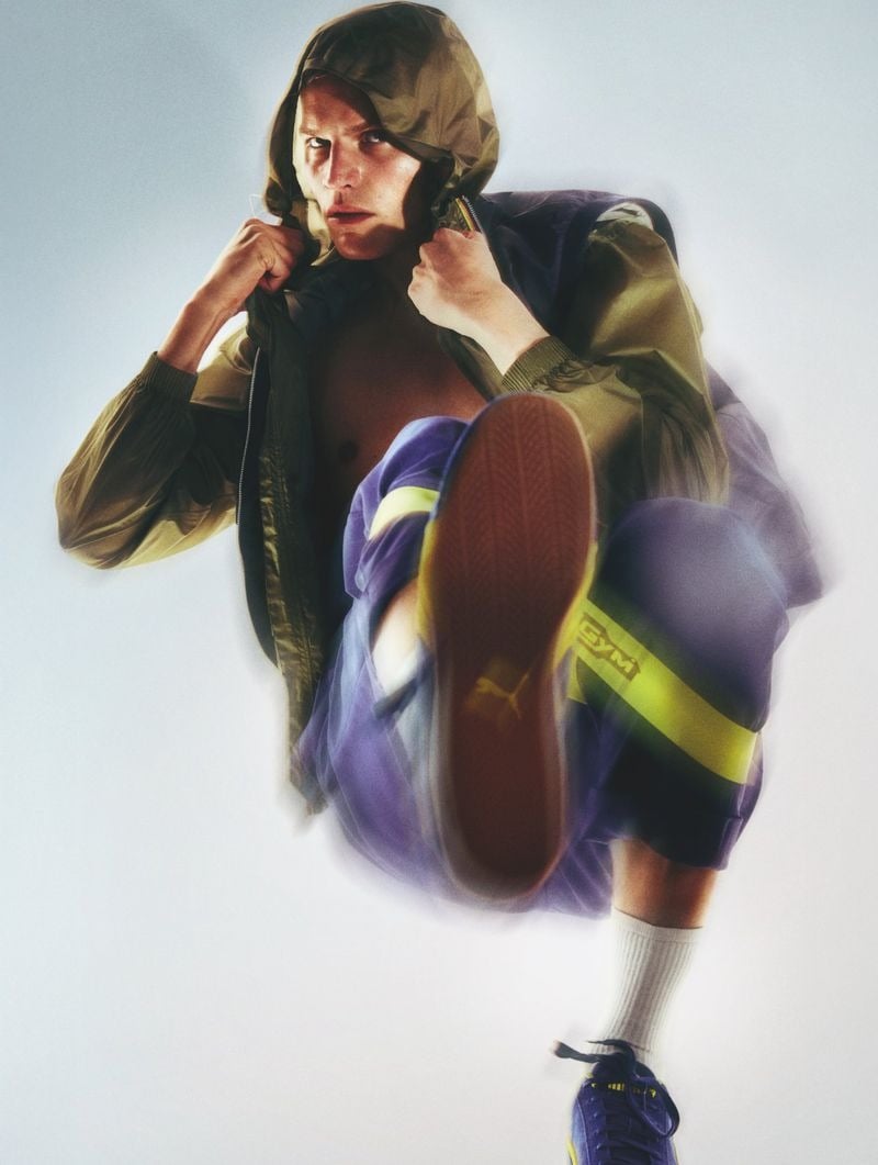 Timo Pan pulling the hood of an olive khaki nylon zip windbreaker tight around his face, wearing lilac purple shorts with a yellow side stripe, white crew socks, and navy Puma 'GYM' sneakers, one raised foot turned to camera with motion blur against a white backdrop