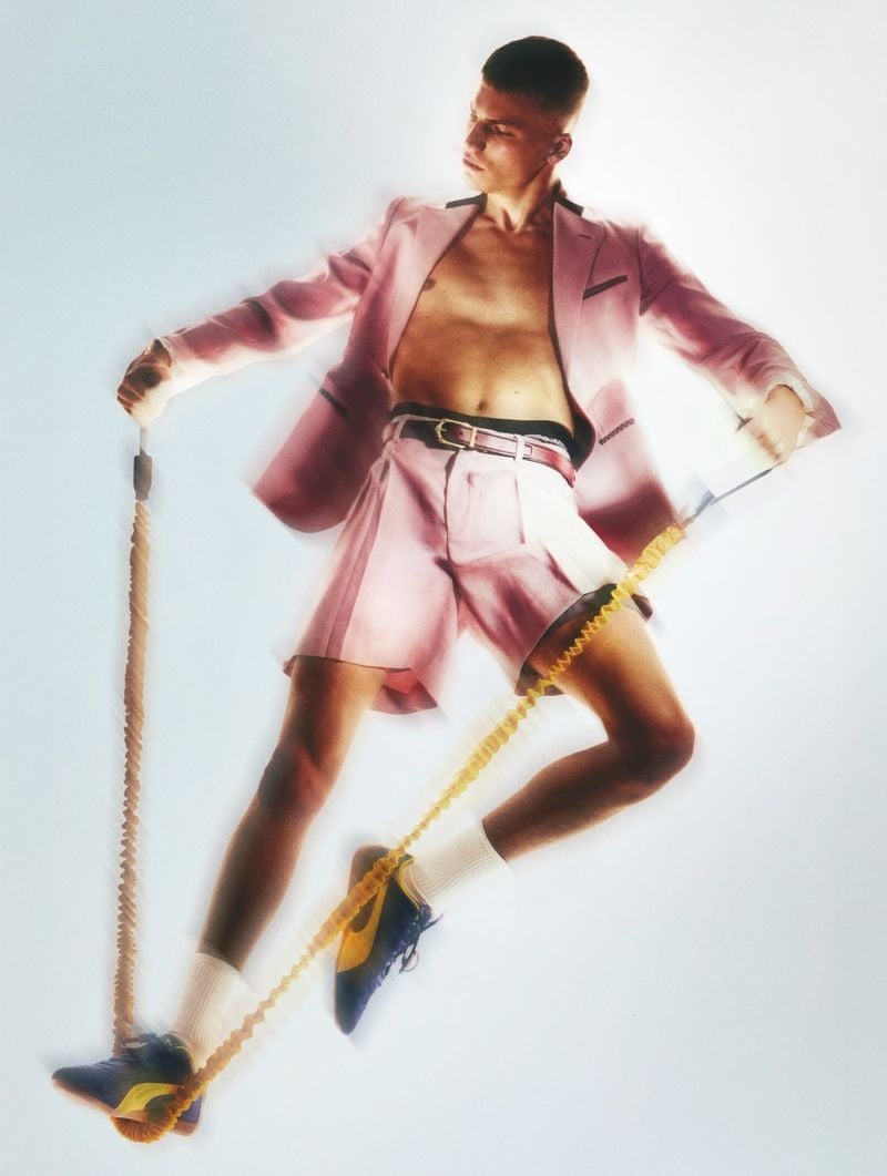 Full-length shot of Timo Pan from the cover in a pink Louis Vuitton blazer over bare chest, matching pleated pink shorts with a brown belt, white tube socks and blue Puma suede sneakers, stretching a yellow Technogym resistance bungee between his hands and feet against a white backdrop