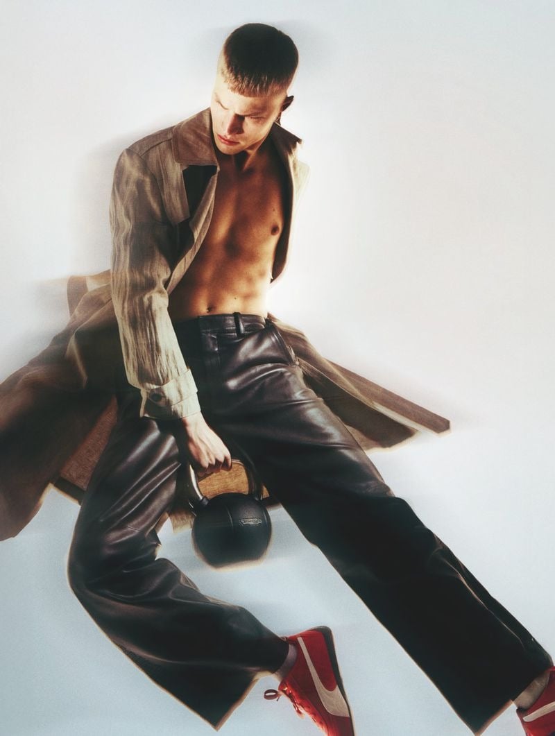 High-angle shot of Timo Pan reclining shirtless on a white floor in an open tan linen trench coat and high-waisted black leather trousers, holding a black Technogym kettlebell between his legs and a red and white sneaker turned to the camera.