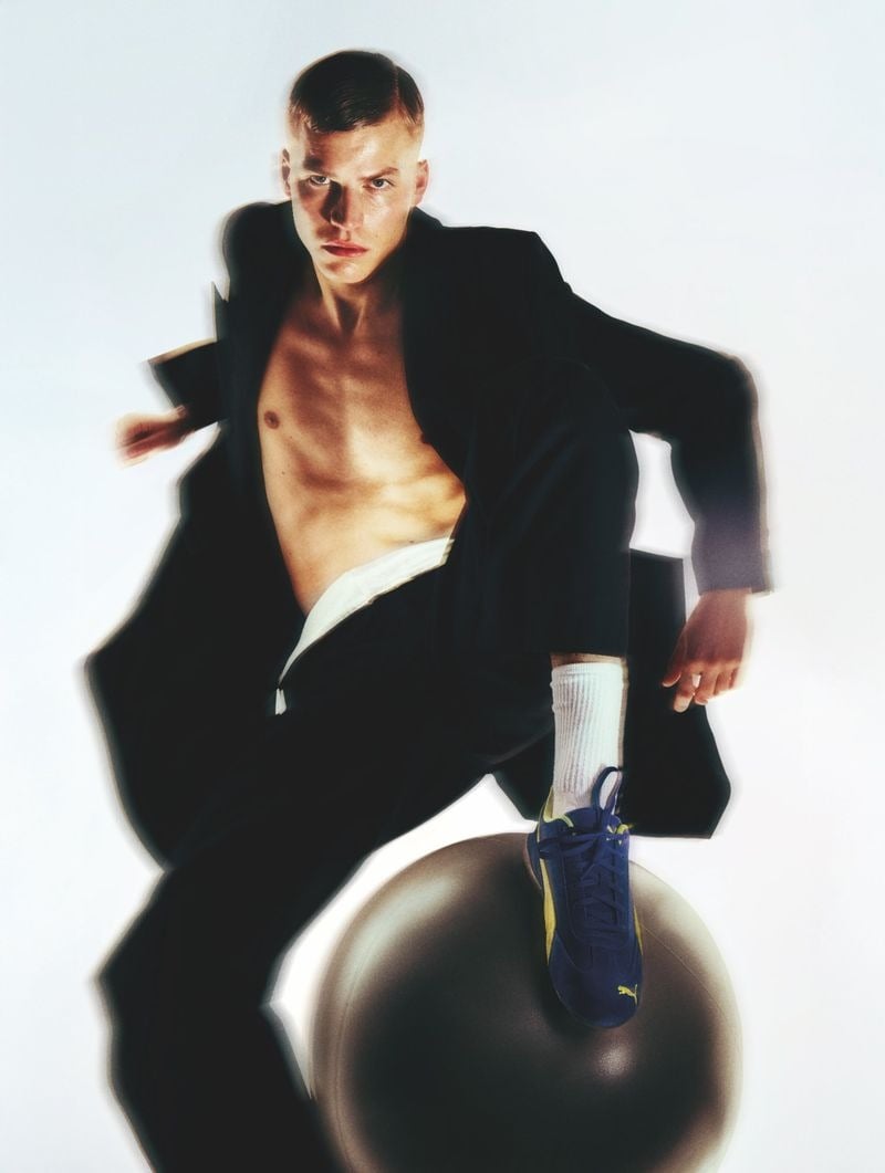 Timo Pan in an oversized black Dries Van Noten blazer thrown open across a bare chest with matching black trousers and white briefs visible at the waist, one foot in a navy and yellow Puma suede sneaker balanced on a gray Technogym exercise ball against a white backdrop