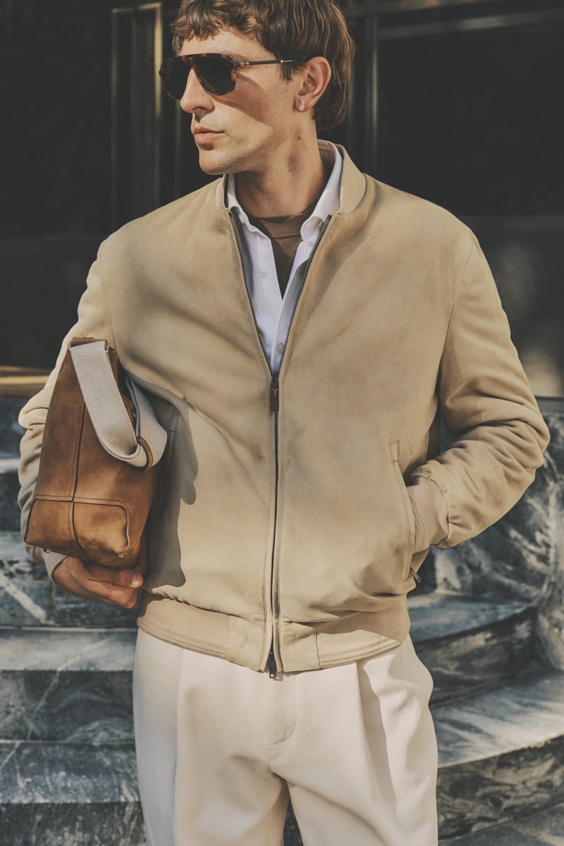 Model Joshua (Tomorrow is Another Day) in tortoise aviator sunglasses wears a sand suede bomber jacket over an olive knit polo and white shirt, paired with cream pleated trousers and a tan leather tote