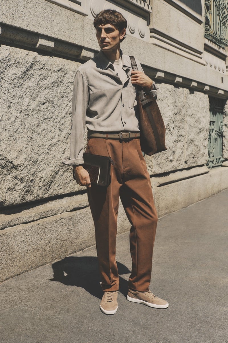 Model Joshua (Tomorrow is Another Day) in a sand suede shirt over a tee, paired with tobacco brown pleated trousers, a brown braided belt, and sand suede sneakers, carrying a chocolate brown suede tote