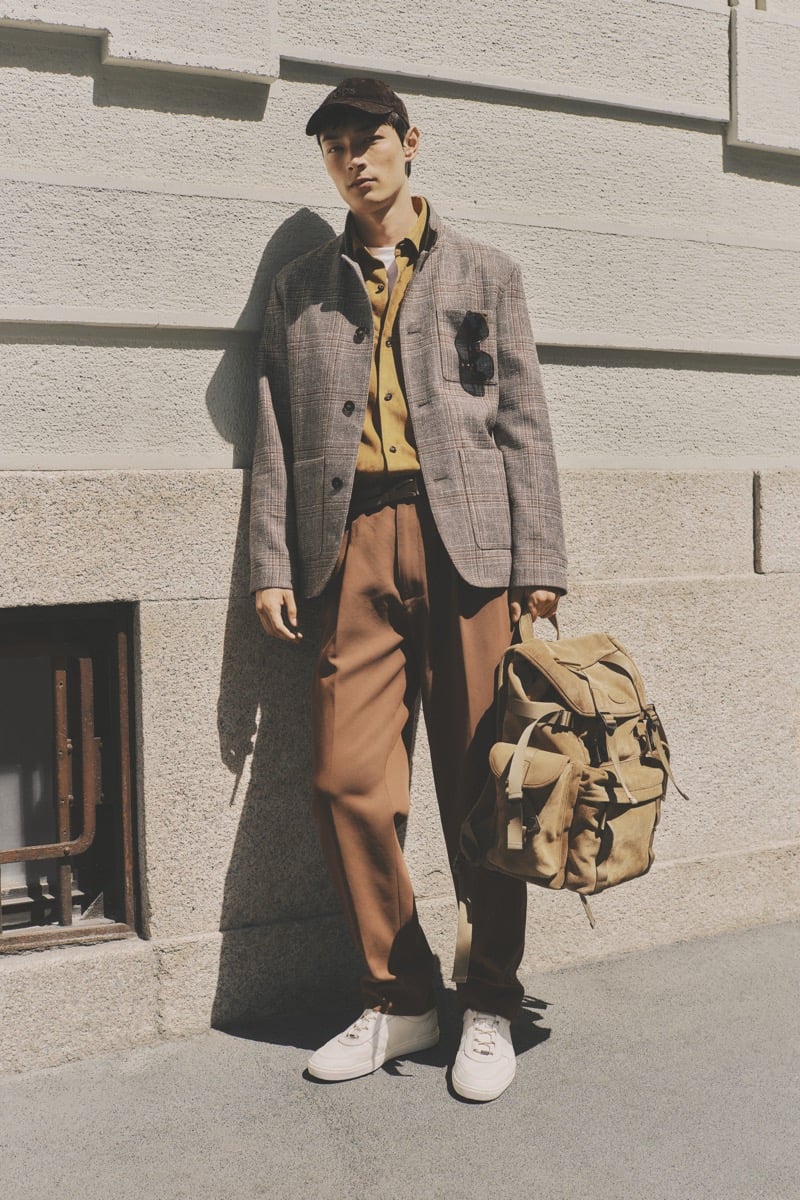 Model Pascal Thulin in a brown corduroy cap wears a gray Prince of Wales check blazer over a mustard yellow shirt and white tee, paired with brick brown pleated trousers and white sneakers, holding a tan canvas and leather backpack
