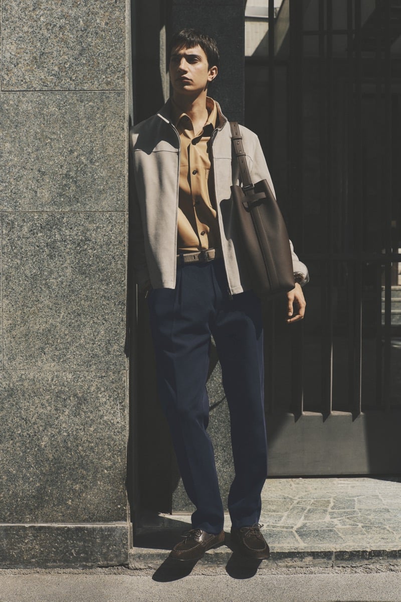 Model Alejandro Albaladejo in a stone suede bomber jacket over a camel button-down shirt, paired with navy pleated trousers, brown loafers, and a taupe leather messenger bag